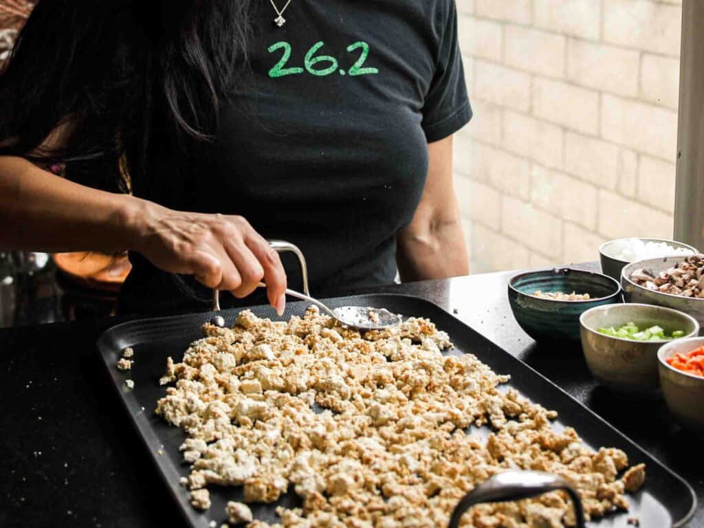 A person wearing a black shirt with 26.2 in green cooks crumbled tofu on a griddle, with bowls of chopped vegetables placed nearby on a countertop.